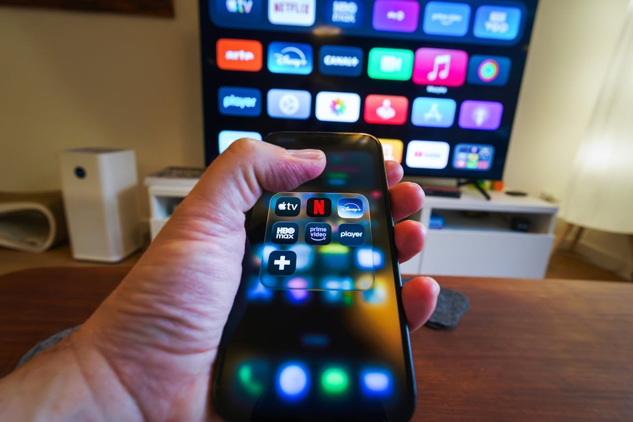hero-img-02 Close-up of a hand holding a phone displaying streaming apps in front of a TV with multiple app icons.